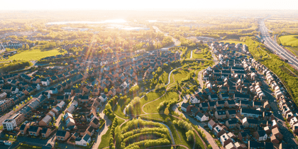 Aerial view of suburban housing development at sunset with winding green spaces, lakes, and highway in the distance.