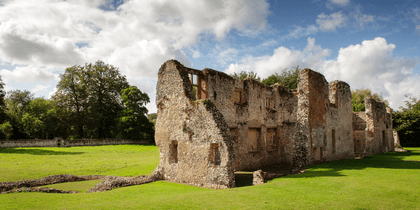 Ancient stone ruins of a medieval building with crumbling walls standing on green grass under a partly cloudy blue sky.